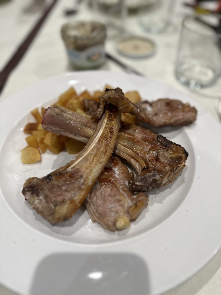 Fotografía de un plato de chuletas de cordero con patatas en Casa Perú, Bagergue. Las chuletas están doradas y jugosas, y las patatas están cortadas en rodajas y cocinadas hasta quedar tiernas. El plato está servido en un plato de cerámica rústico.