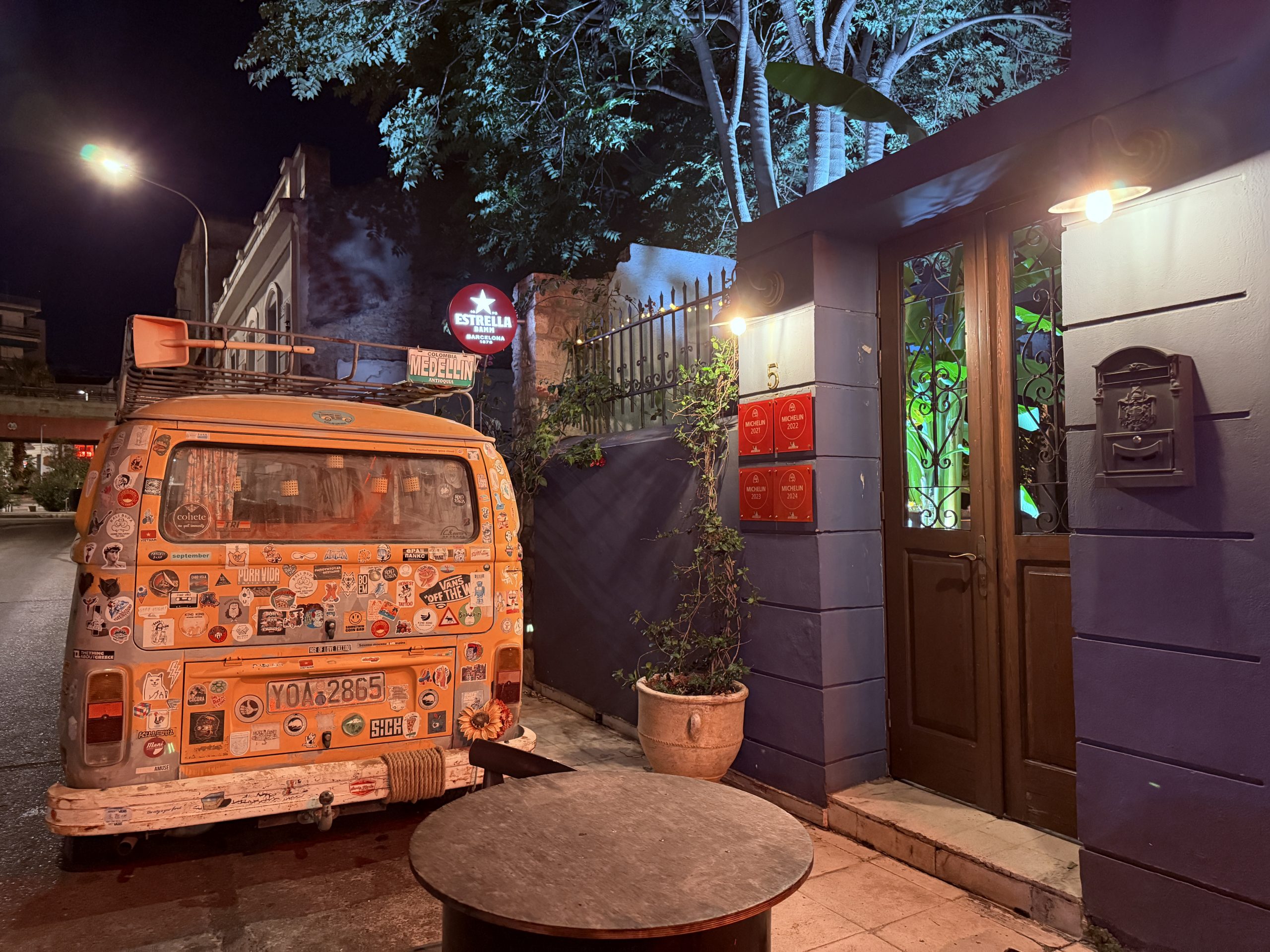 Entrada del restaurante El Cerdo Negro 1985 en Atenas, con varias menciones Michelin visibles en rojo y una furgoneta estilo surf aparcada en la puerta