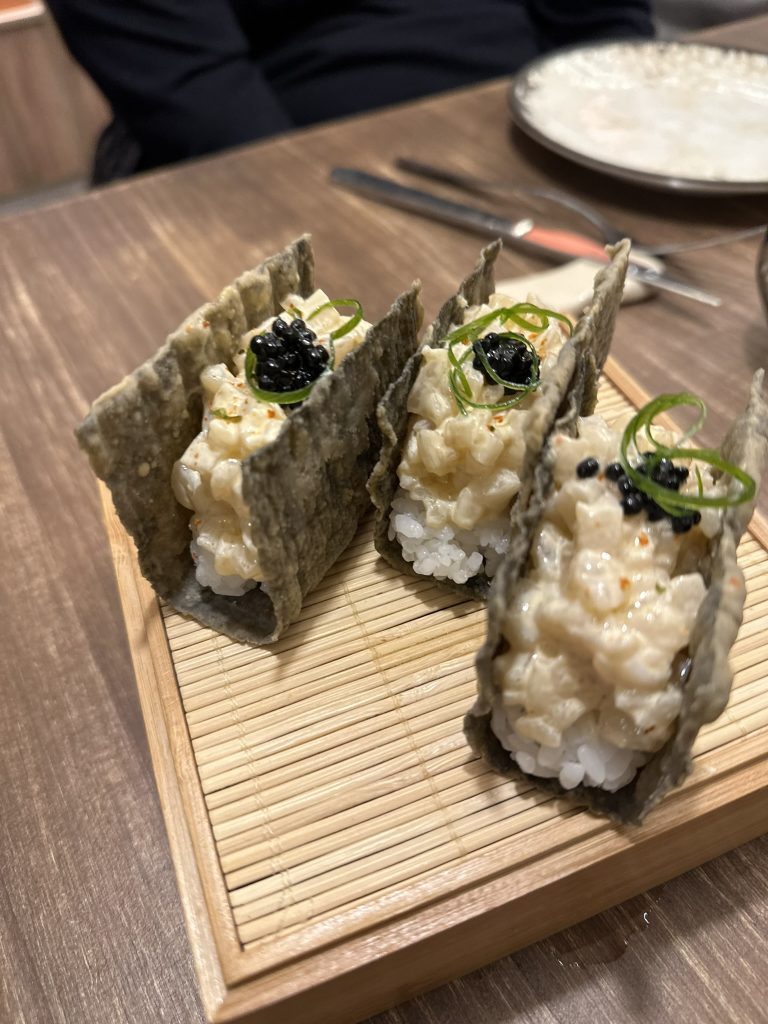 Un taco crujiente de alga nori relleno de tartar de vieira, arroz de sushi y un toque de aceite de trufa.