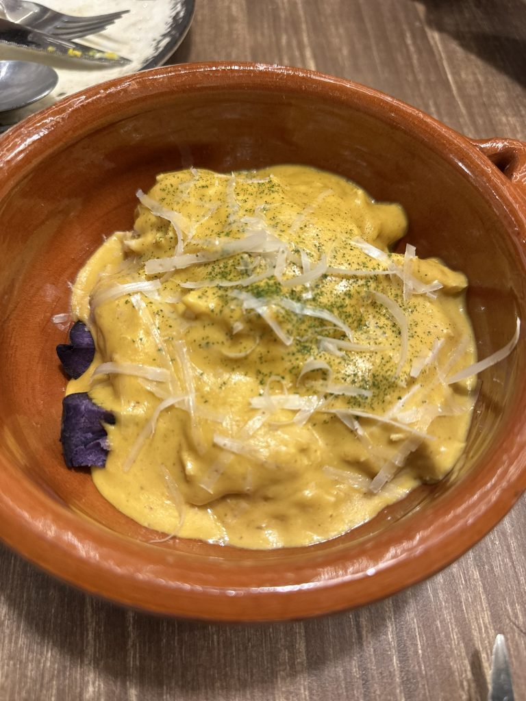 Un plato de ají de gallina con pollo desmenuzado bañado en una cremosa salsa amarilla, acompañado de rodajas de papa violeta y decorado con aceitunas y huevo cocido.