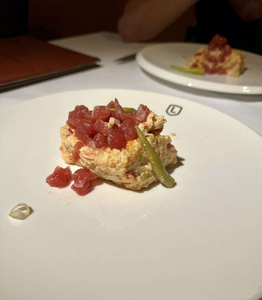Plato blanco con una porción redonda de ensaladilla cremosa coronada por trozos de tartar de atún rojo y varias piparras verdes, sobre un fondo neutro