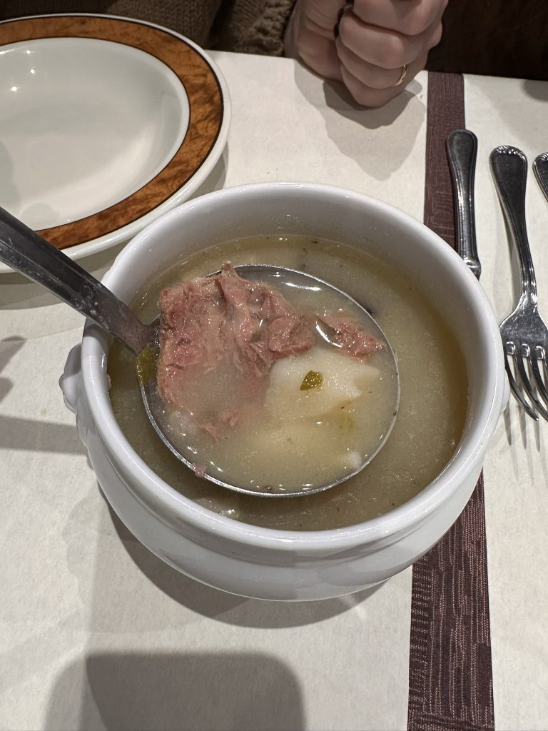 Fotografía de un plato de Olla Aranesa en Casa Perú, Bagergue. La olla, de barro, contiene un caldo caliente con verduras y carne. Se aprecian trozos de patata, zanahoria, col y carne de cerdo. El plato está servido en una mesa de madera rústica.