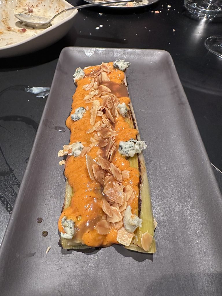 Puerro confitado servido con romescu de sobrasada, queso azul fundido y almendras fritas crujientes.